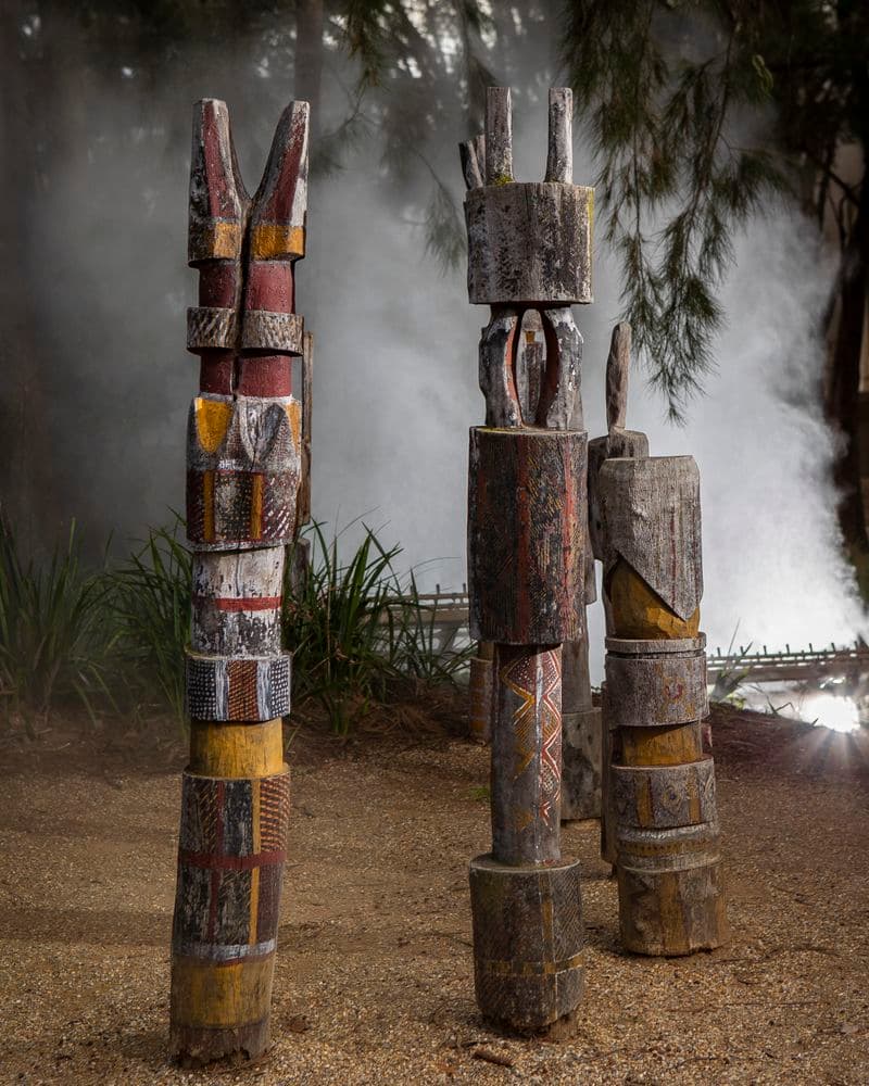 Three carved poles painted in red, yellow, and white ochre with mist behind