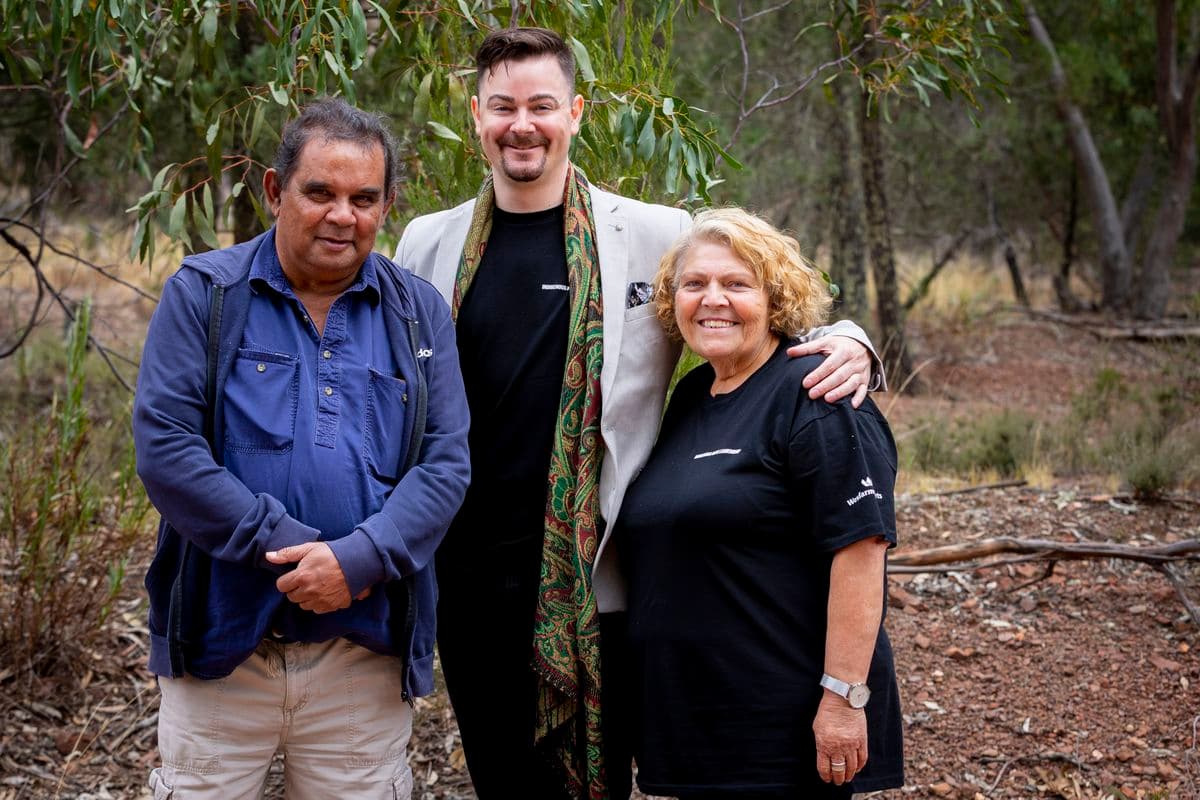Photo of two men and a woman are standing outside beneath a gum tree. The man in the middle has his arms around the others, while they look at the camera smiling.