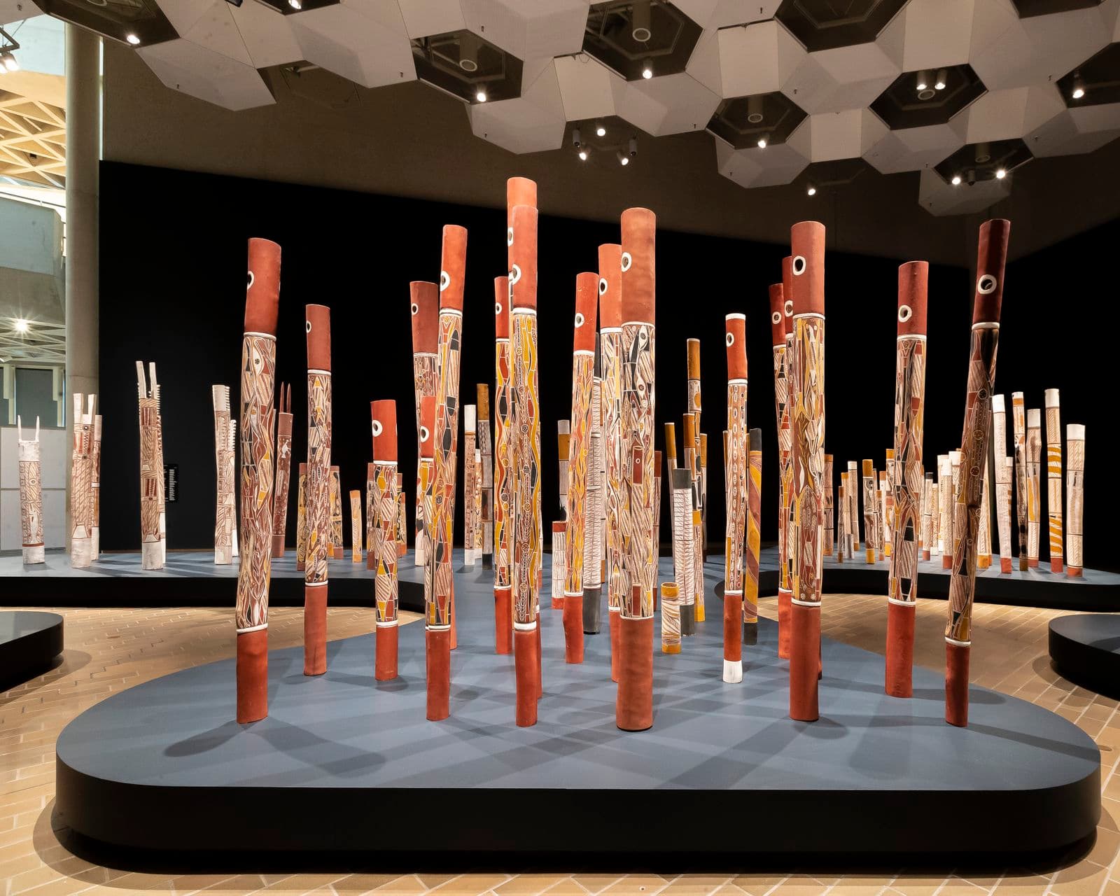 A photograph showing a section of an installation of 200 painted hollow logs standing upright in a large gallery space. The surrounding walls are painted in a dark colour and the logs are positioned on a navy plinth shaped to resemble both sides of a river bank.