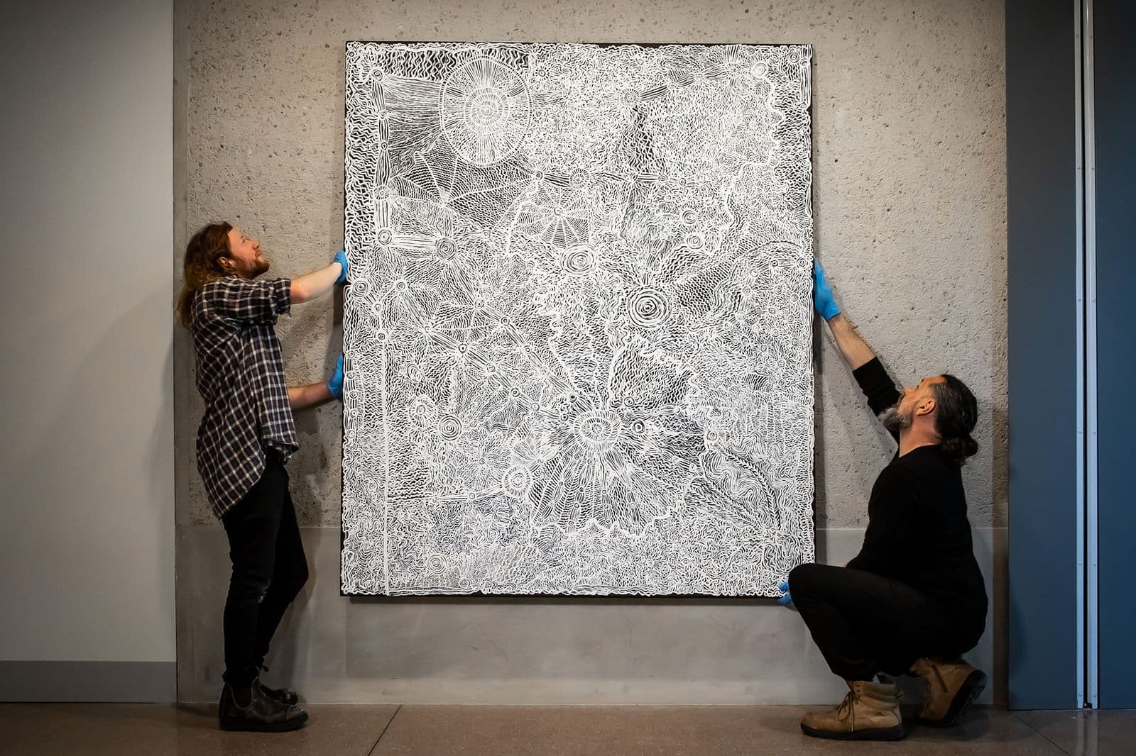 two men wearing gloves are hanging a large canvas on a concrete wall