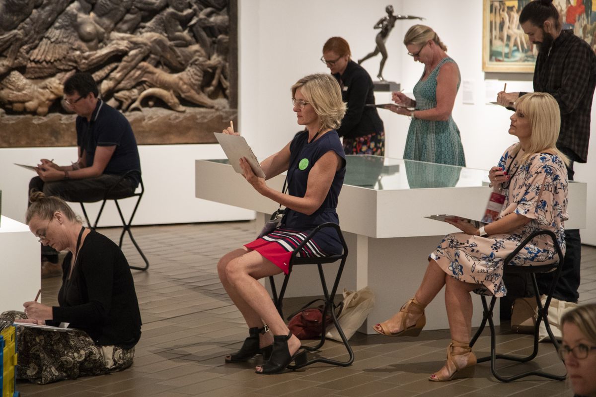 A group of people are seated facing a group of artworks sketching them