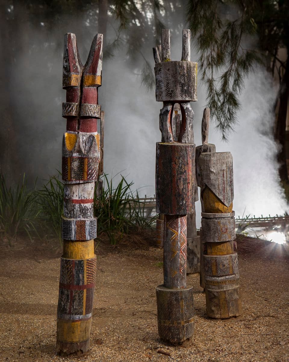 Three carved poles painted in red, yellow, and white ochre with mist behind