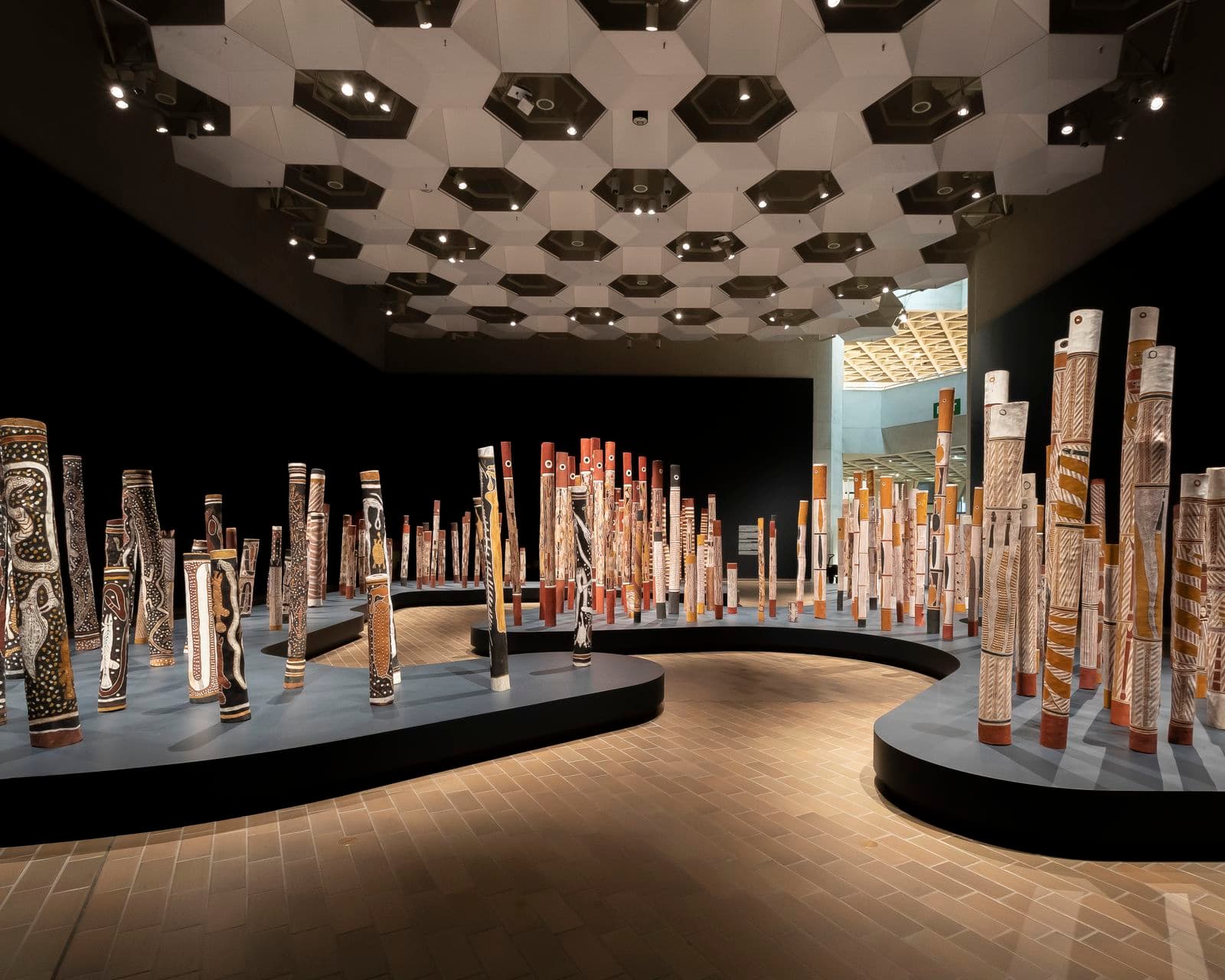 A wide-angle photograph showing an installation of 200 painted hollow logs standing upright in a large gallery space. The surrounding walls are painted in a dark colour and the logs are positioned on a navy plinth shaped to resemble both sides of a river bank.