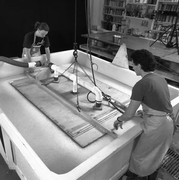 Black and white image of two men lowering the Frank Stella ‘Moby Dick Domes’ shaped papermaking vacuum mould into the vat of paper pulp, while vacuum suction pulls the pulp slurry against the submerged mould,
