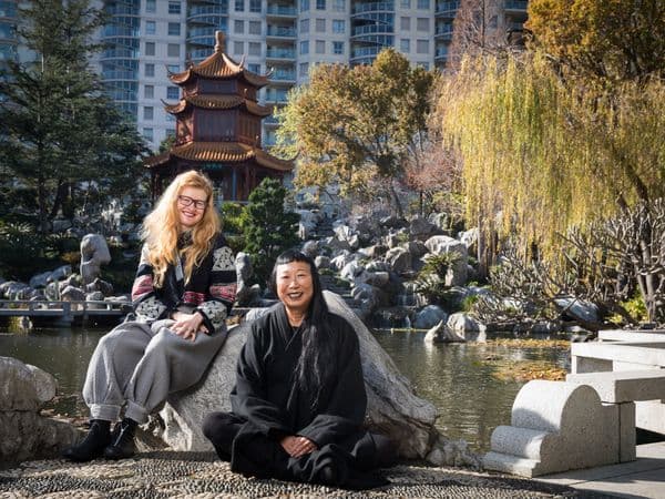Artist Nell sitting on a rock next to artist Lindy Lee sitting cross-legged on the ground in a Chinese garden