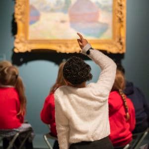 Photograph of school children sitting in front of painting with arms raised to answer question