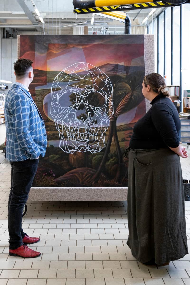 A man and a woman are standing on either side of a work of art that is sitting on an a-frame trolley, looking towards it. They are inside a conservation lab.
