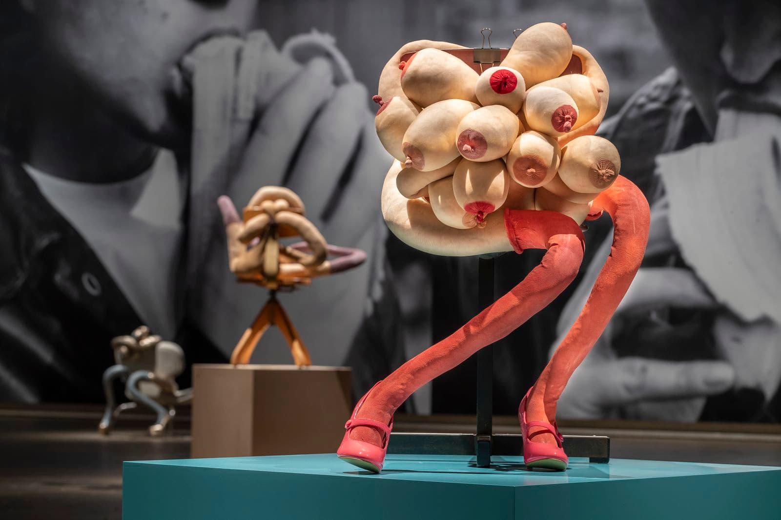 Installation view of a sculpture of a human-like form made up of a cluster of many breasts and two long legs with long pink socks, sitting on a chair