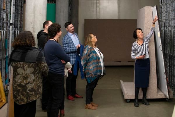 Five people are watching a demonstration given by a woman in a painting storage area.