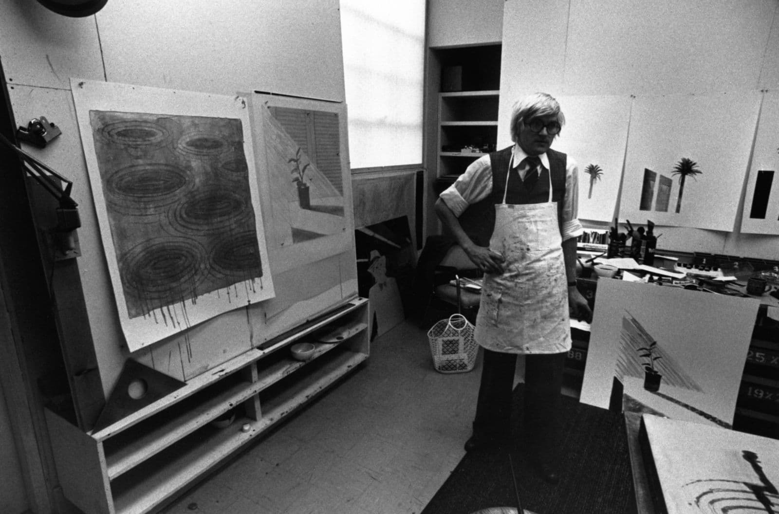 The artist David Hockney, sporting a golden bowl of straw-like hair, and the thickest of rimmed glasses, stands neutrally in the middle of a room, wearing an apron. He is surrounded by lithographs of climactic conditions that he has encountered in Los Angeles.