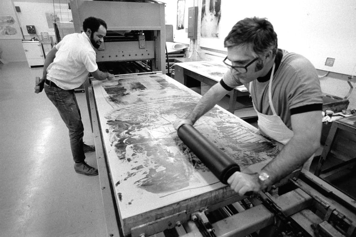 Two men in an art workshop rolling ink onto a large rectangular stone