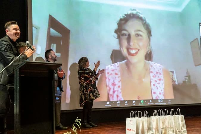 Three people are on stage clapping, while a large projection on the stage of a video call shows a person on screen smiling