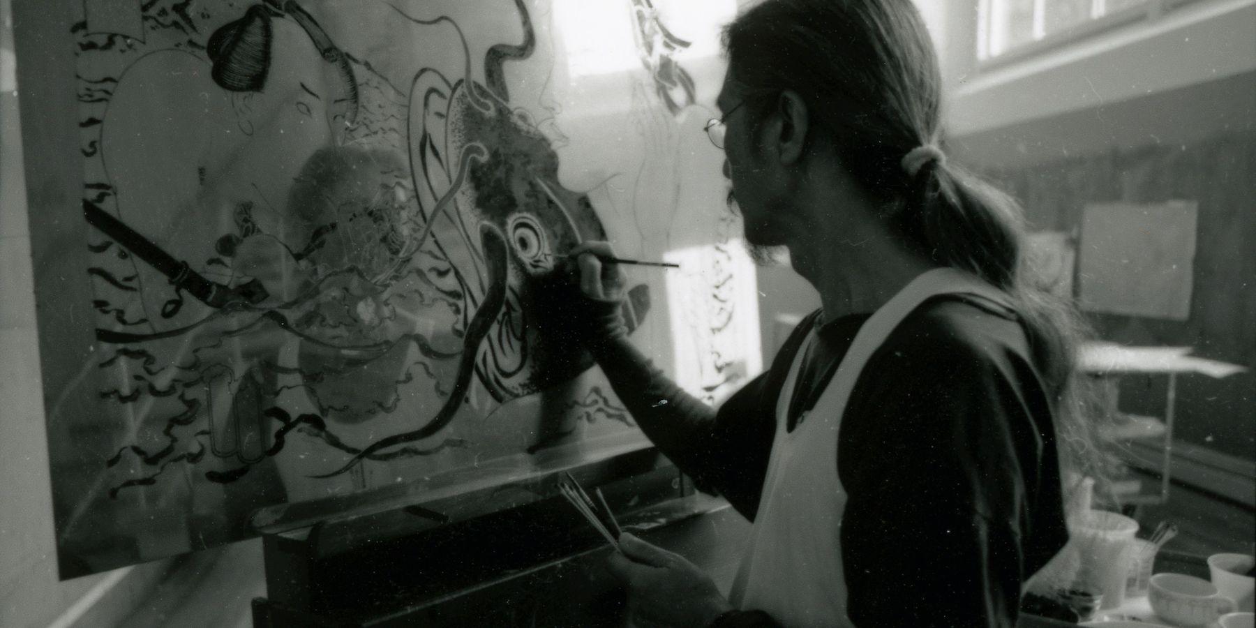 A blurry black and white photograph of Masami Teraoka painting.