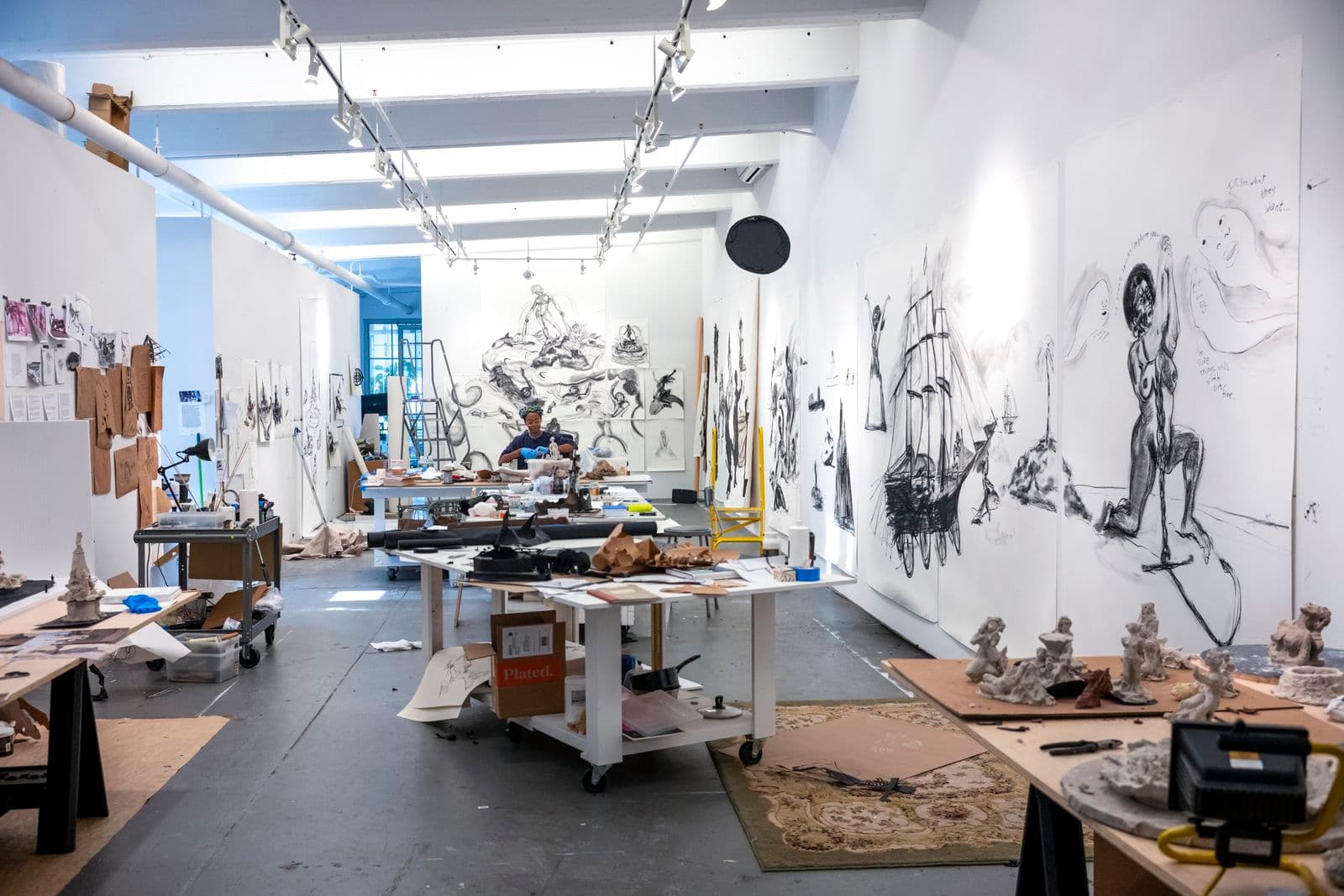An artist's studio with works on paper pinned to the wall and tables of mixed media. An artist is working in the background with gloves handling clay.