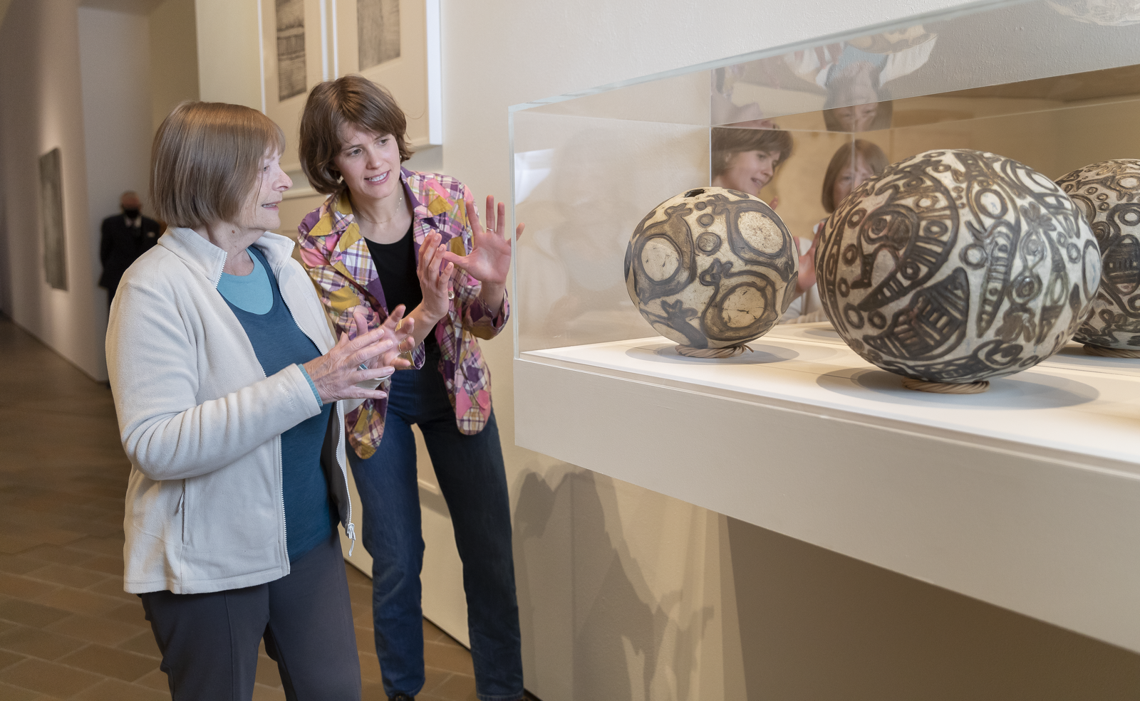 Photograph of two women standing by  and gesturing to case with round objects