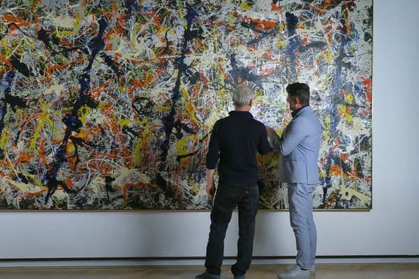Video still of two men looking at painting.