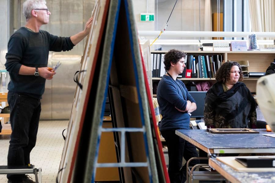 In a conservation lab one man is standing on a footstool and treating a paining resting on an a-frame stand. Nearby, two other people are watching a demonstration out of frame..