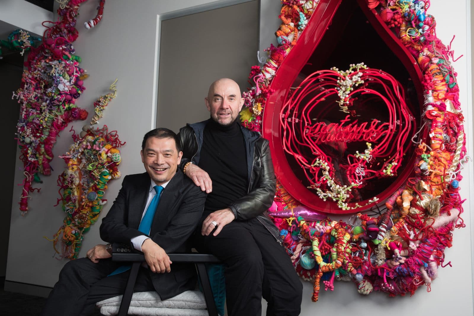 Two men sit together in front of an artwork