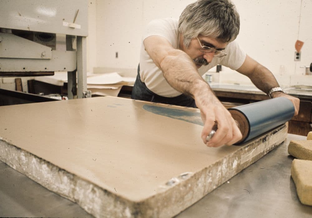 Man in an art workshop using a large paint roller to press paint ink on stone