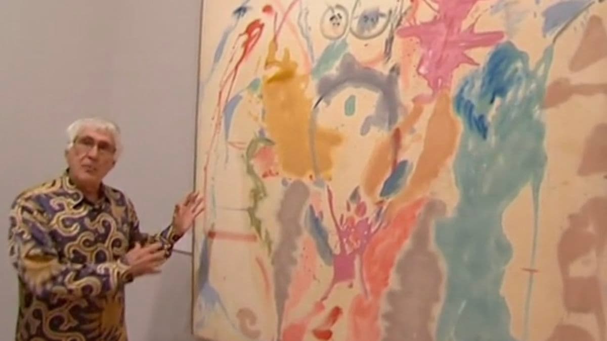 Video still of man talking about painting, that is hanging on wall beside him.
