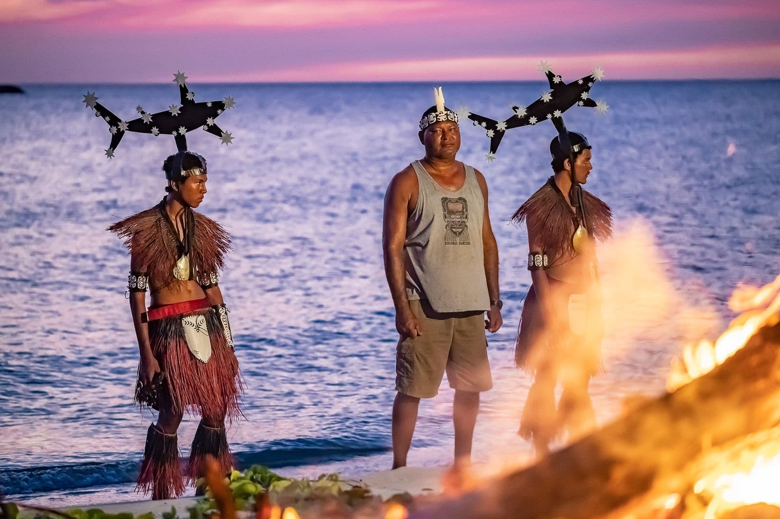 Photograph of artist Alick Tipoti standing on a beach on Badhu Island, in the Zenadth Kes/Torres Straits at sunset with other members of his community is ceremony dresss