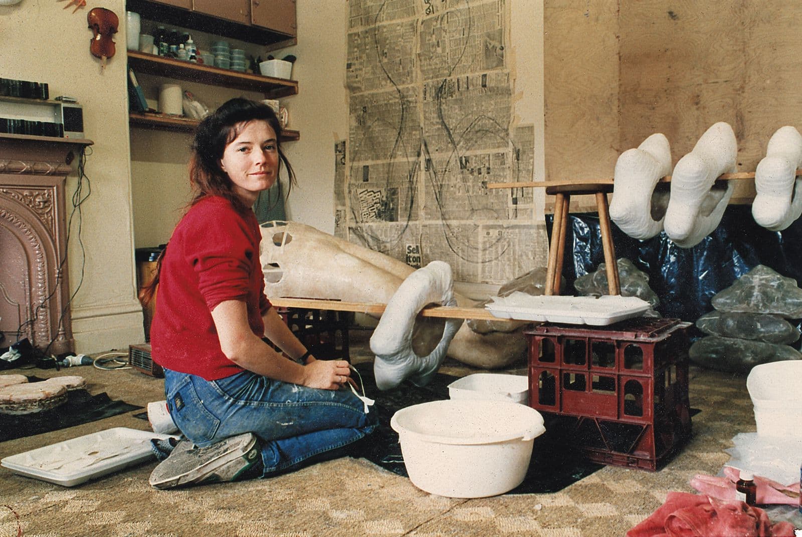 A photograph of a woman in a red shirt gazing back at the camera in an artists studio.