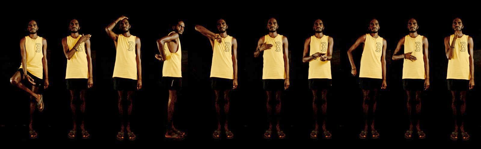 A wide long photograph / still from a film with a First Nations man, wearing a yellow singlet and black shorts signing
