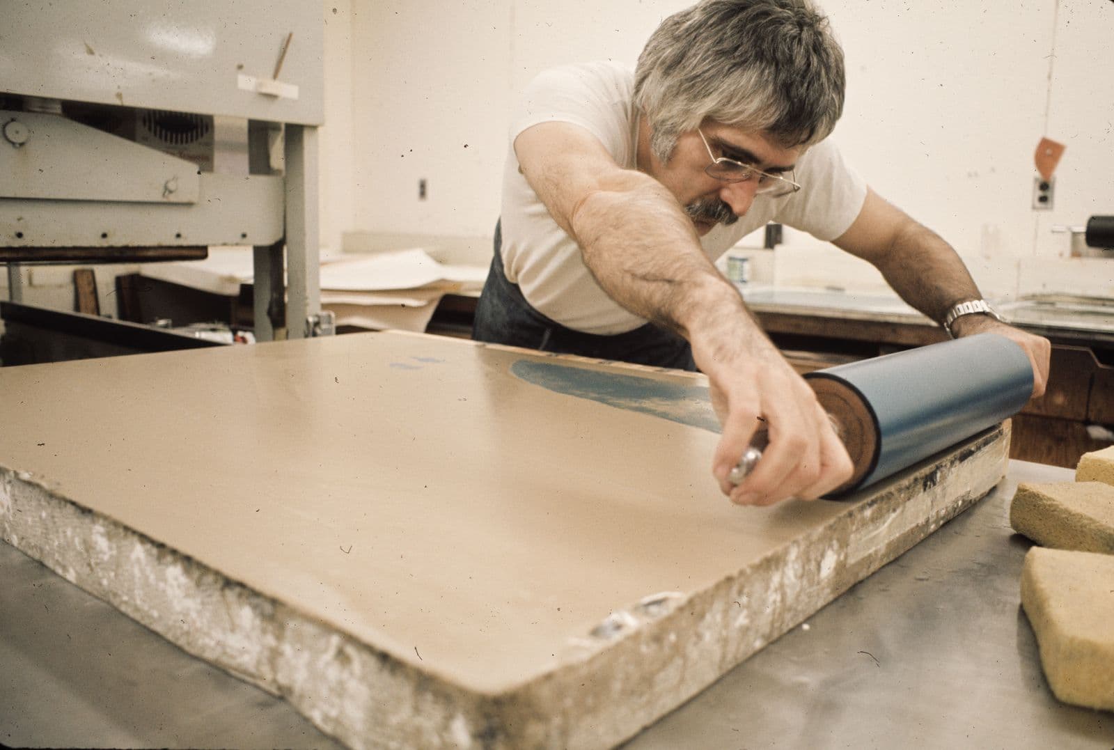 Man in an art workshop using a large paint roller to press paint ink on stone