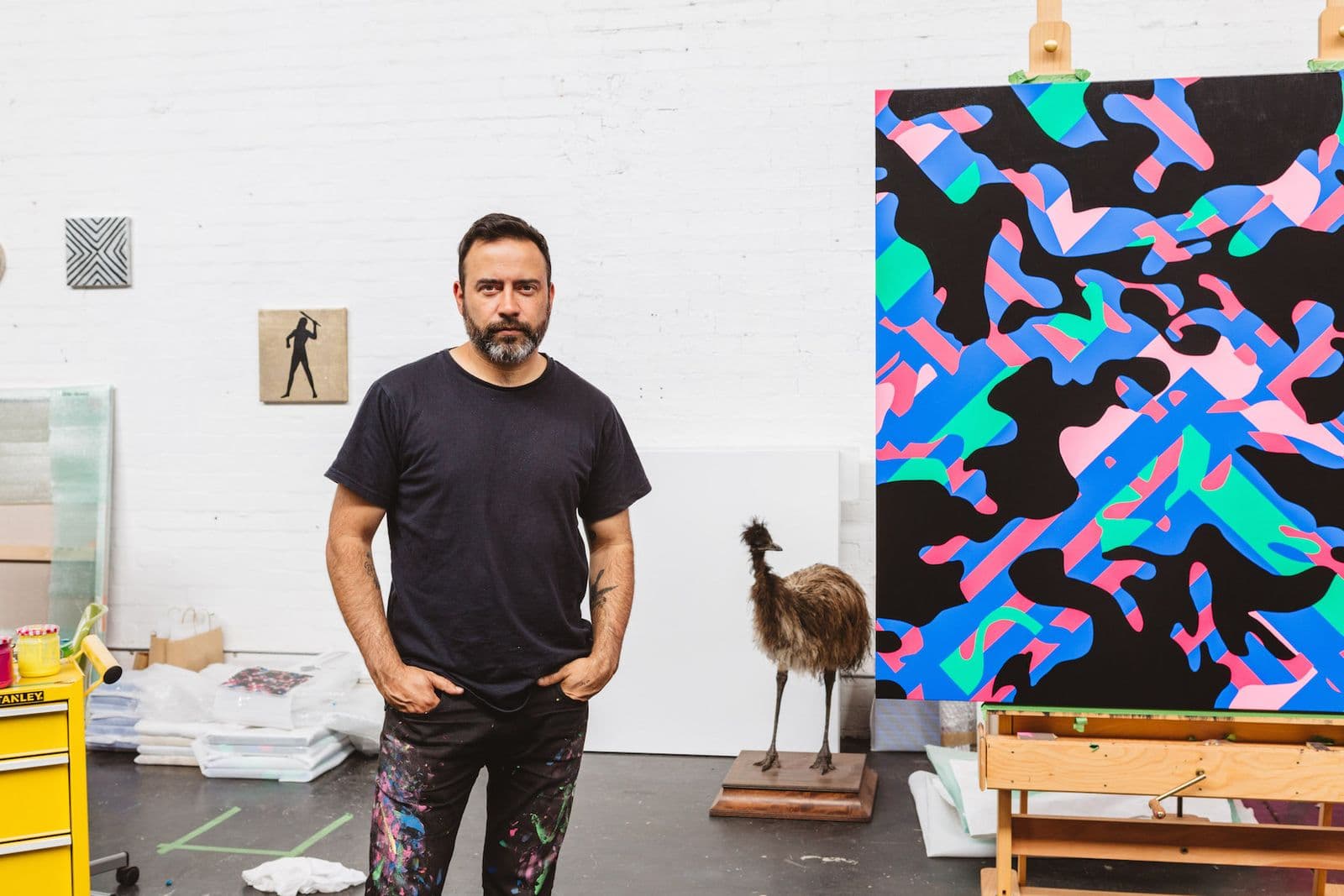 An image of Reko Rennie in his studio in a black shirt with his hands in his pockets.