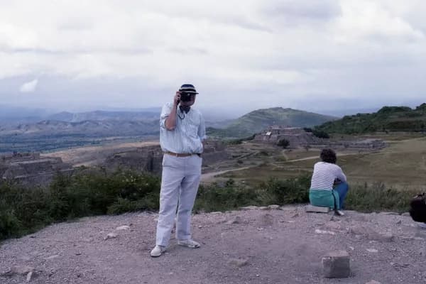 Colour photograph of David Hockney holding a camera to his eye in front of Oaxaca ruins