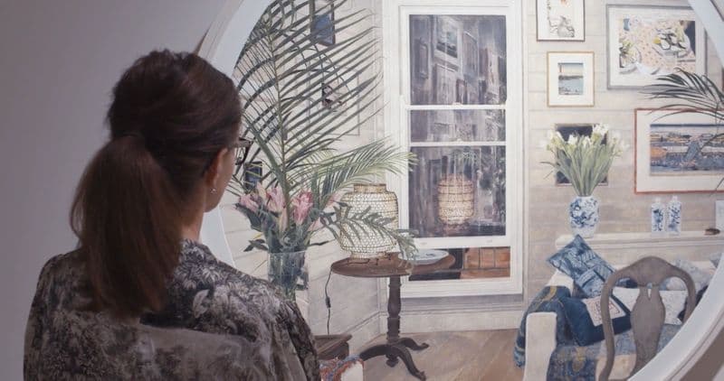 Video of still of the back of a woman wearing a floral pattern shirt with hair in ponytail, looking at a work of art in a gallery of an home interior scene
