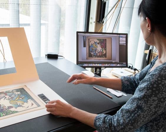 A woman sits at a desk cross-checking a work of art on paper with an image on a computer screen