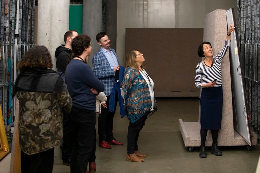 Five people are watching a demonstration given by a woman in a painting storage area.