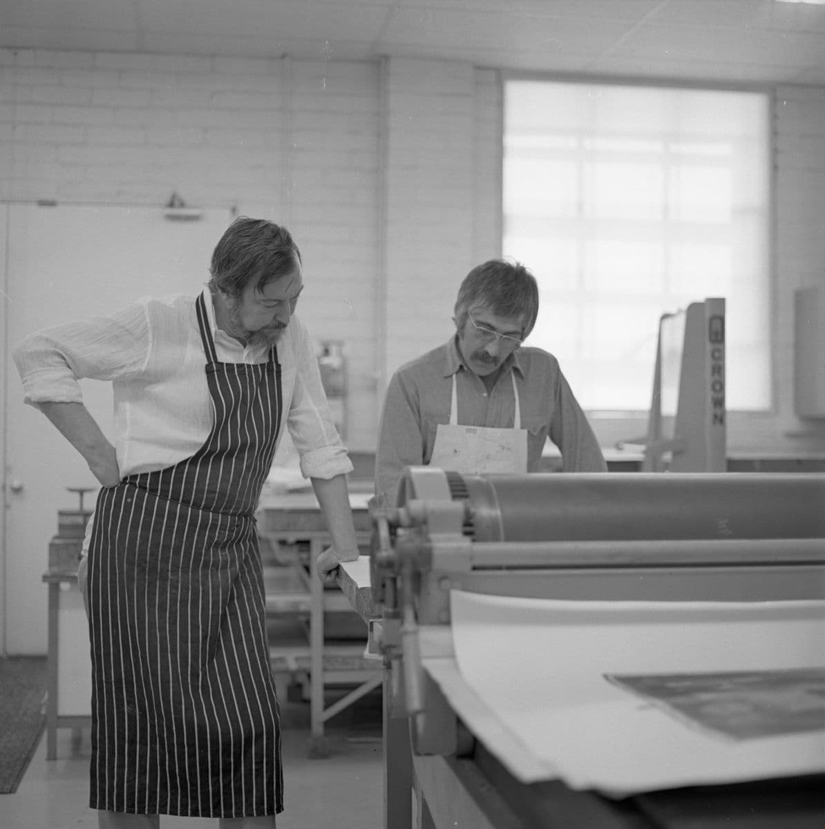 A black and white image of two men in a printmaking studio wearing aprons and inspecting a work