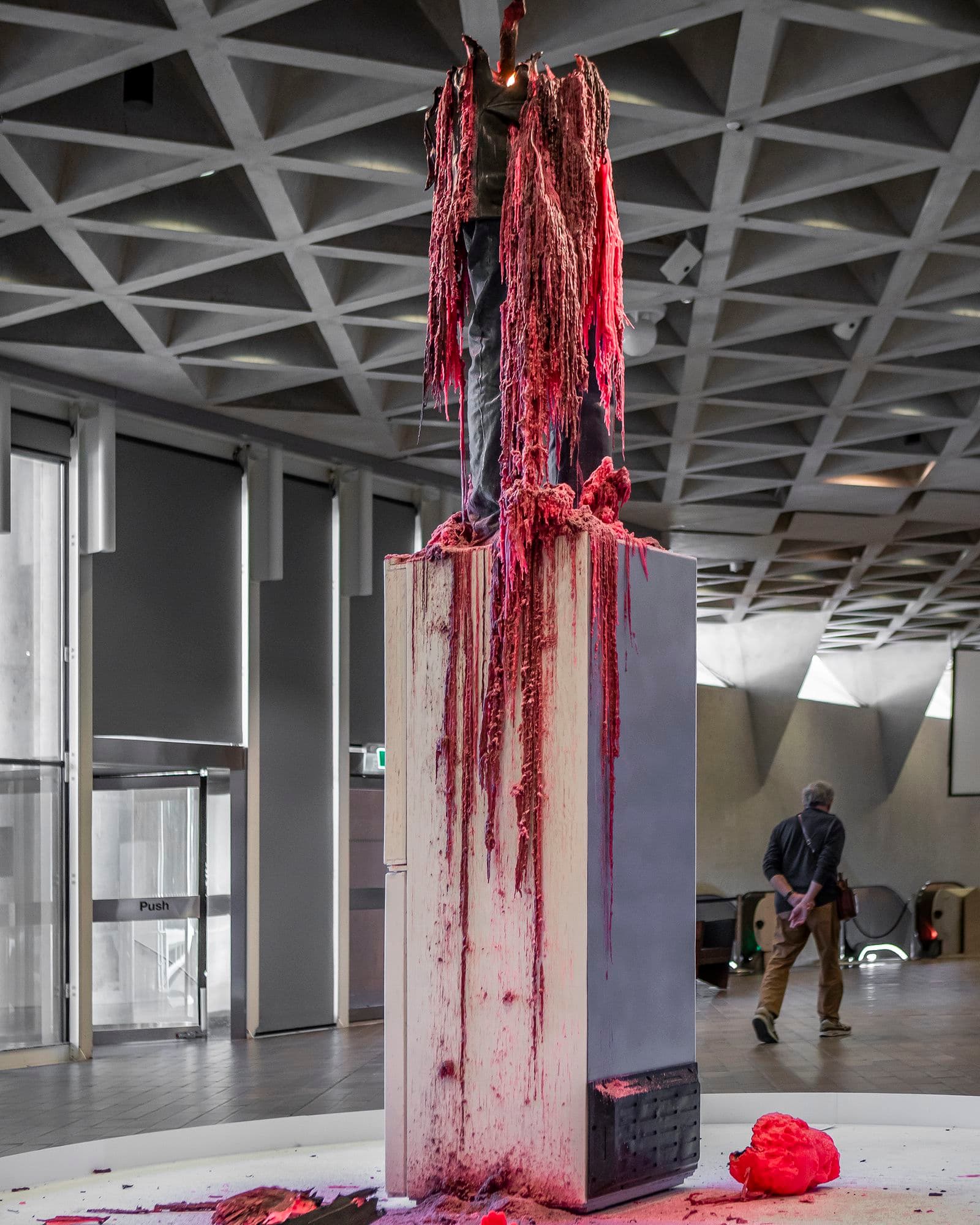 a tall wax sculpture of a man standing upon a fridge is half-melted, with pink and grey wax dripping  down onto the fridge and floor