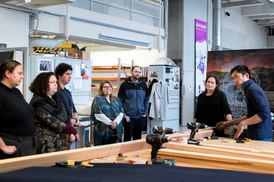 A group of people are standing around large table covered in pieces of timber, drills and other tools, as a man gestures towards the table. He is explaining how to make frames.