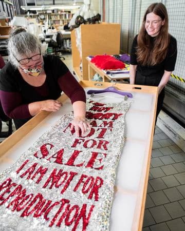 Two women examine a sequinned garment laying flat on a table in a conservation lab