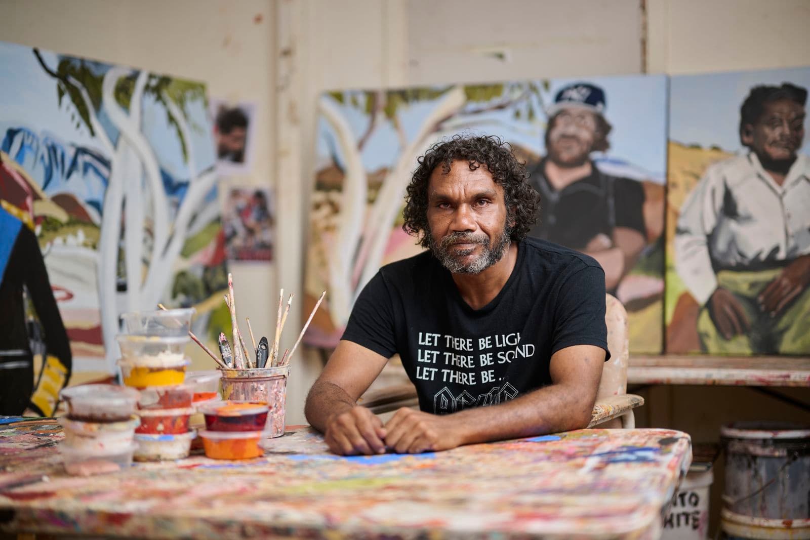 A picture of artist Vincent Namatjira in a studio painting next to a table covered in paints
