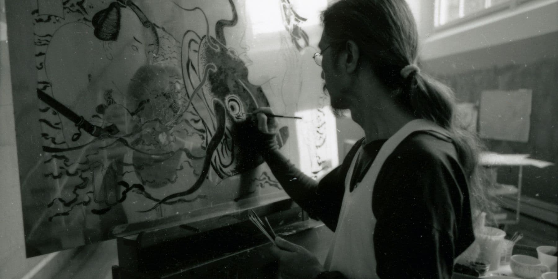 A blurry black and white photograph of Masami Teraoka painting.