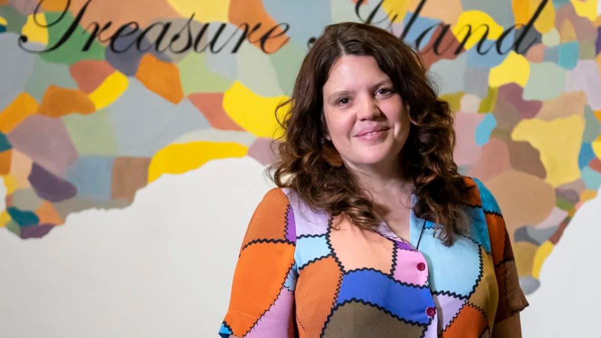 a woman in a bright shirt is smiling and looking at the camera. She is standing in front of a bright painting
