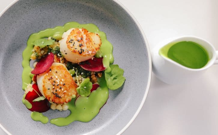 A plate of seared scallops on a bed of vegetables and herbs surrounded by a green sauce