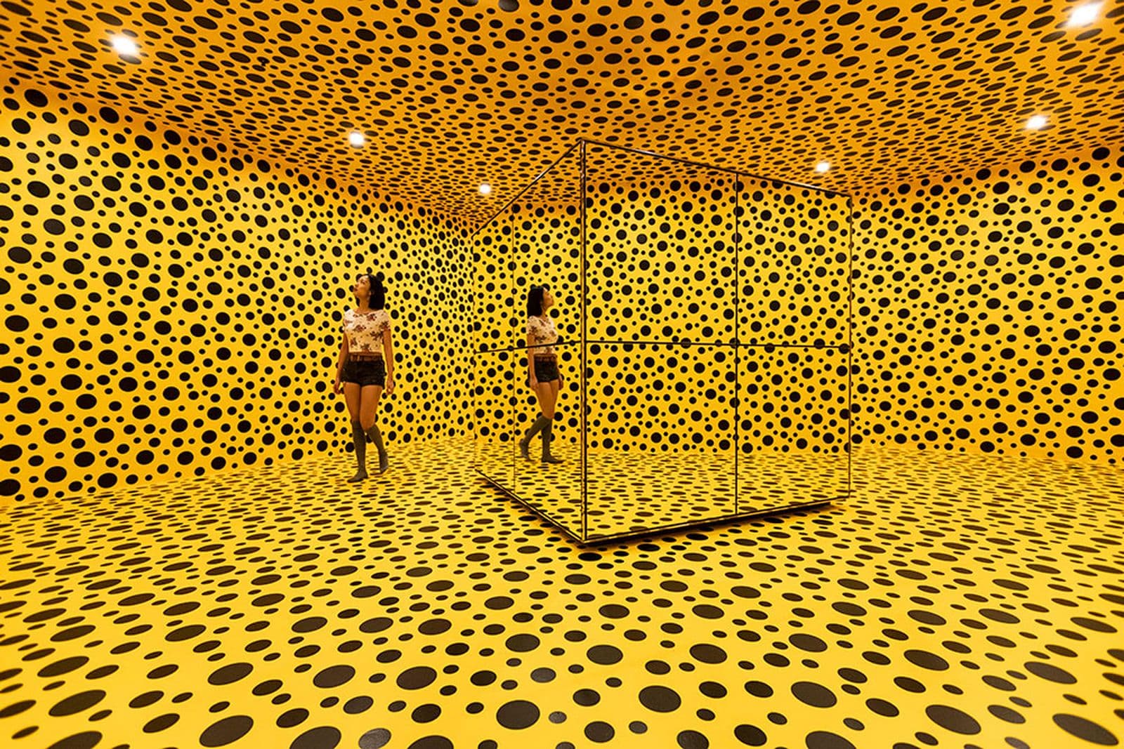 A woman walks through a mirrored yellow room covered in black dots