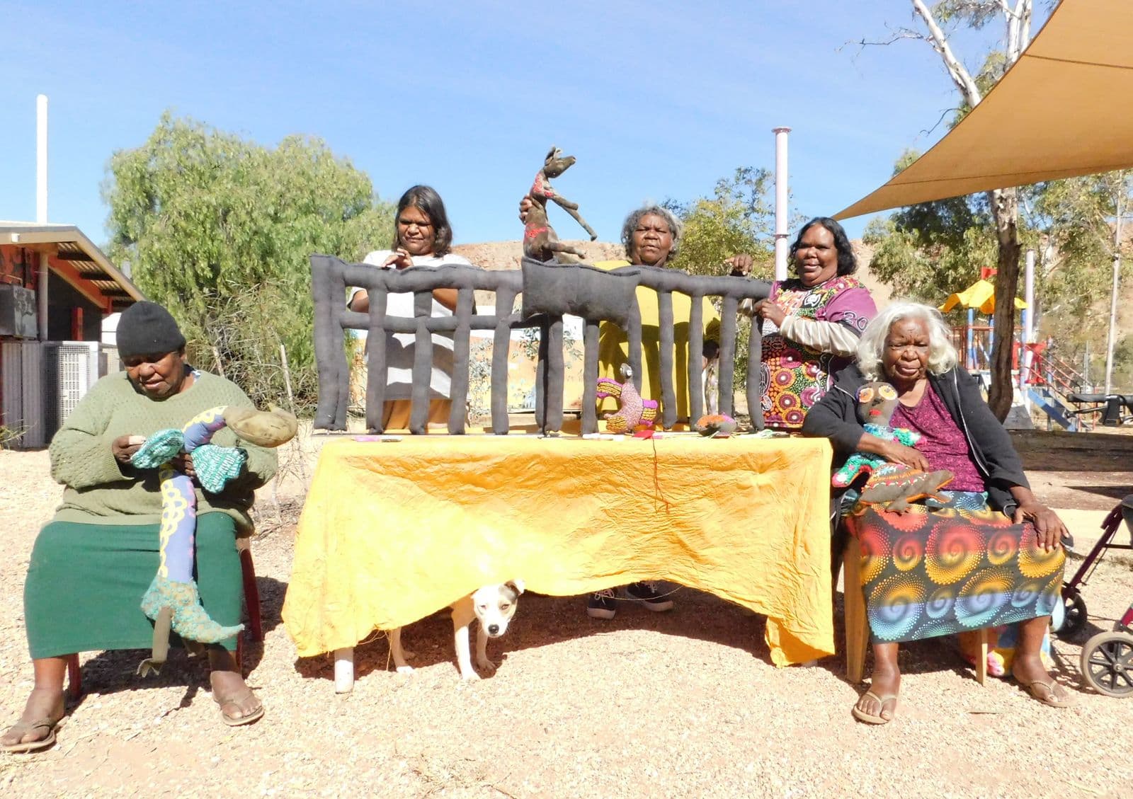 Five Indigenous Australian woman are sitting and standing around a table outside with a bright yellow tablecloth, with a sculpture of parliament house made in black material sitting on the table. A dog is peering out from under the tablecloth.