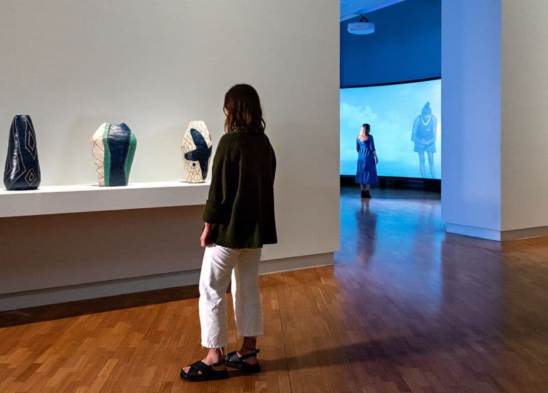 Two people walk through a gallery space looking at various works including vases and a large film