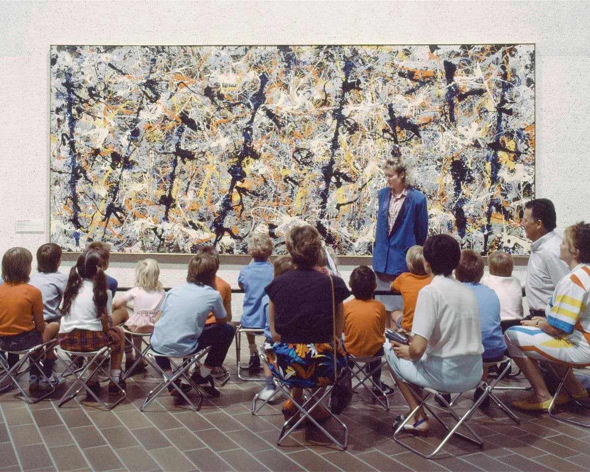 A photograph from the 1980s shows a group of people on stools, sitting in front of Jackson Pollock's Blue poles, with a guide discussing the work.