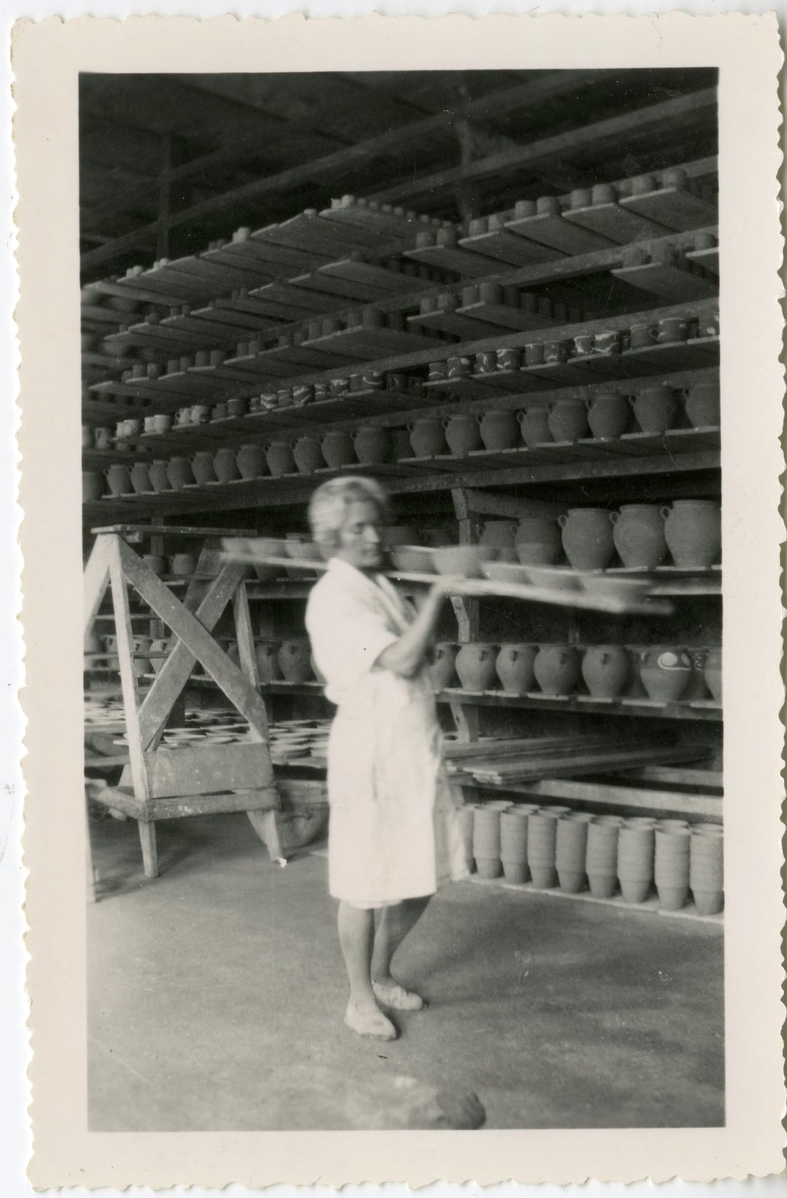 Anne Dangar, in a white dress, carrying a board with a number of unfired, thrown clay bowls. In the background there are shelves full of more vessels.