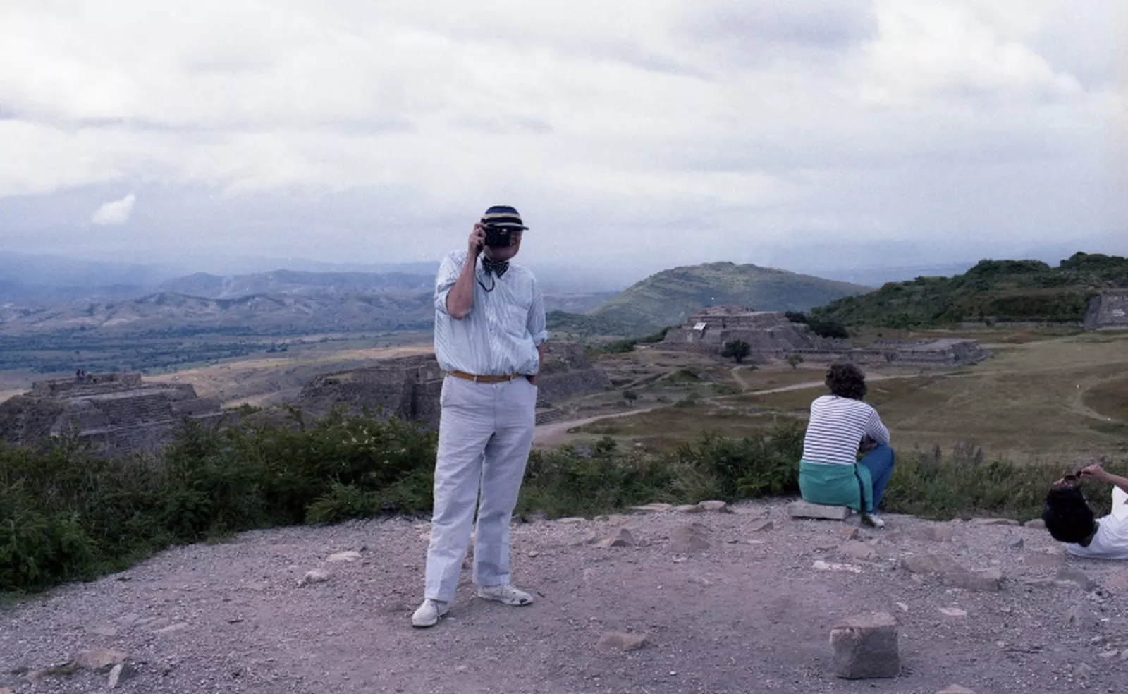 Colour photograph of David Hockney holding a camera to his eye in front of Oaxaca ruins