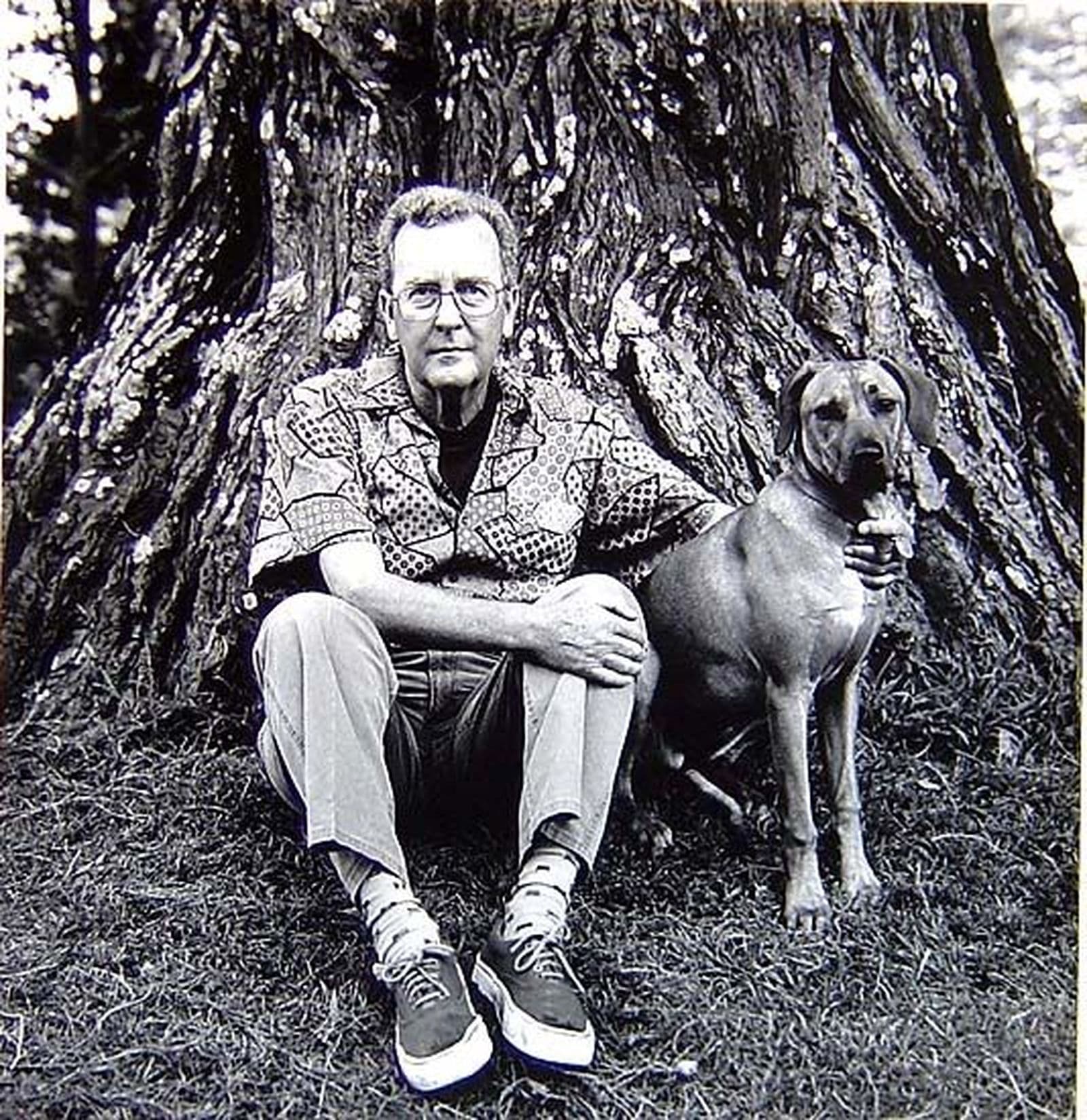 Black and white photograph of man sitting under a tree with a dog.