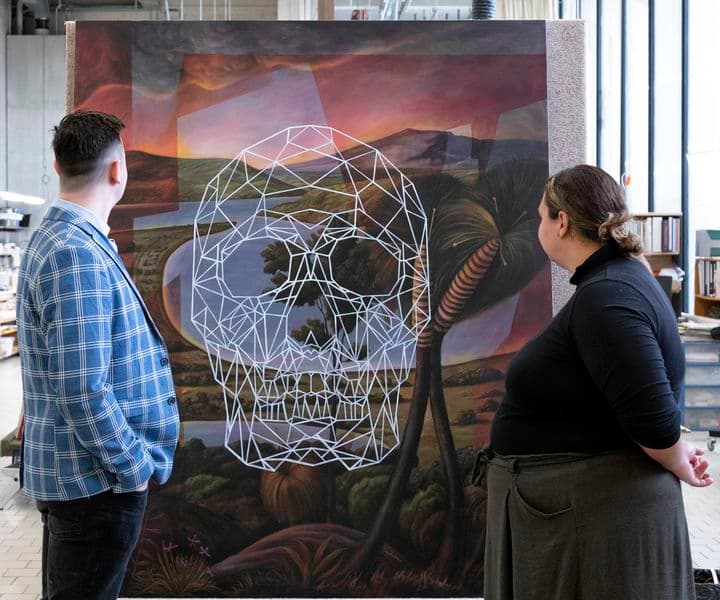 A man and a woman are standing on either side of a work of art that is sitting on an a-frame trolley, looking towards it. They are inside a conservation lab.