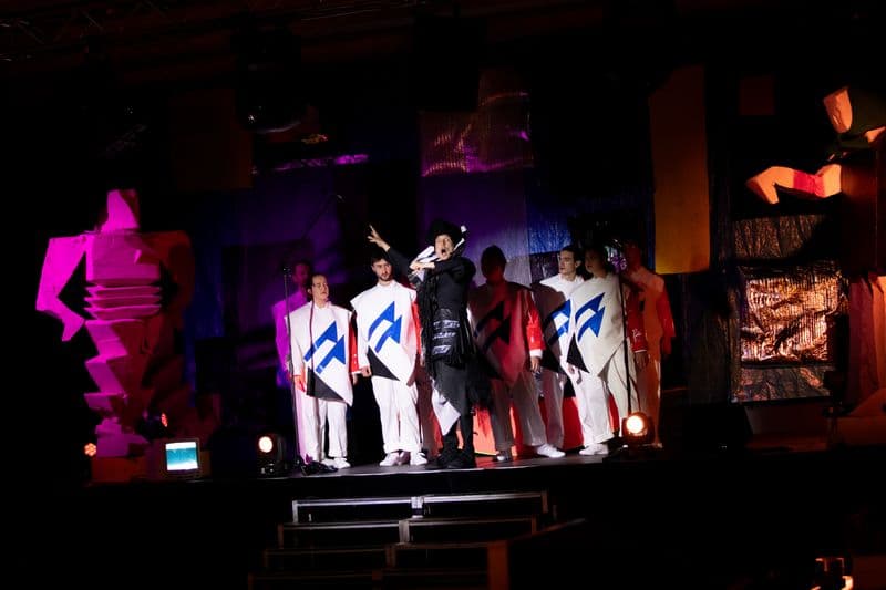 Photo of central performer in elaborate black costume surrounded by choir in white costumes with blue accent